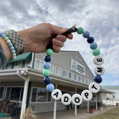 Hand holding a adjustable beaded dog friendship collar that says "ADOPT ME" against a background building of a dog shelter.