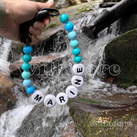 Custom beaded pet collar with blue and white beads and MARINE name letters, highlighted against a natural outdoor waterfall setting for Willadore Collar Co.