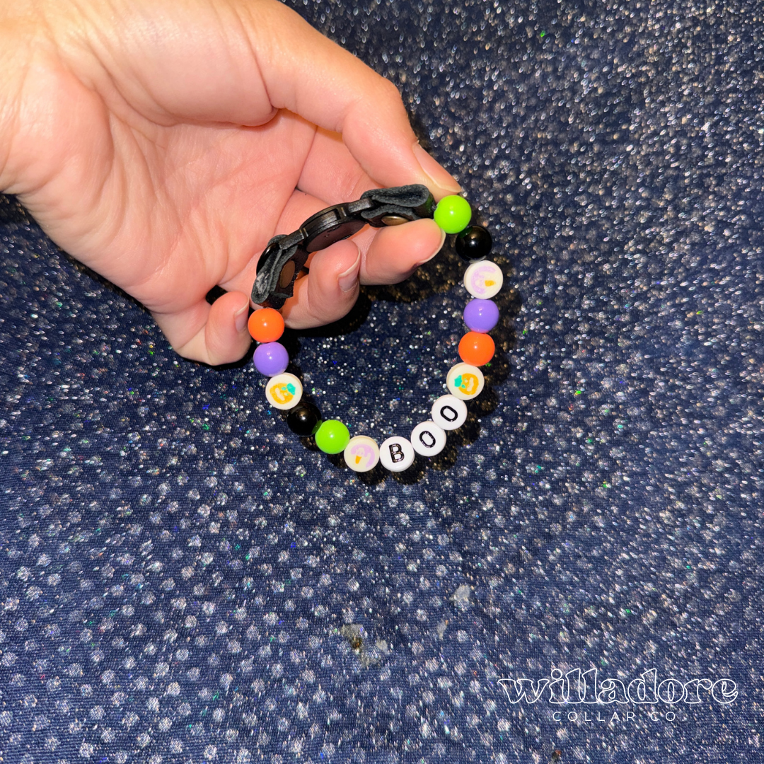 A hand holding a beaded cat collar with colorful Halloween-themed beads and take-and-bake inspired cookie beads, and white letter beads spelling 'BOO', set against a dark blue textured fabric background.